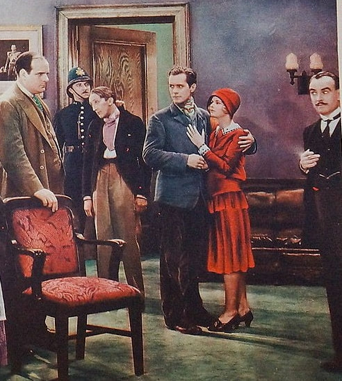Assorted people in a sitting room: a romantic couple, a policeman making an arrest, and two well-dressed men